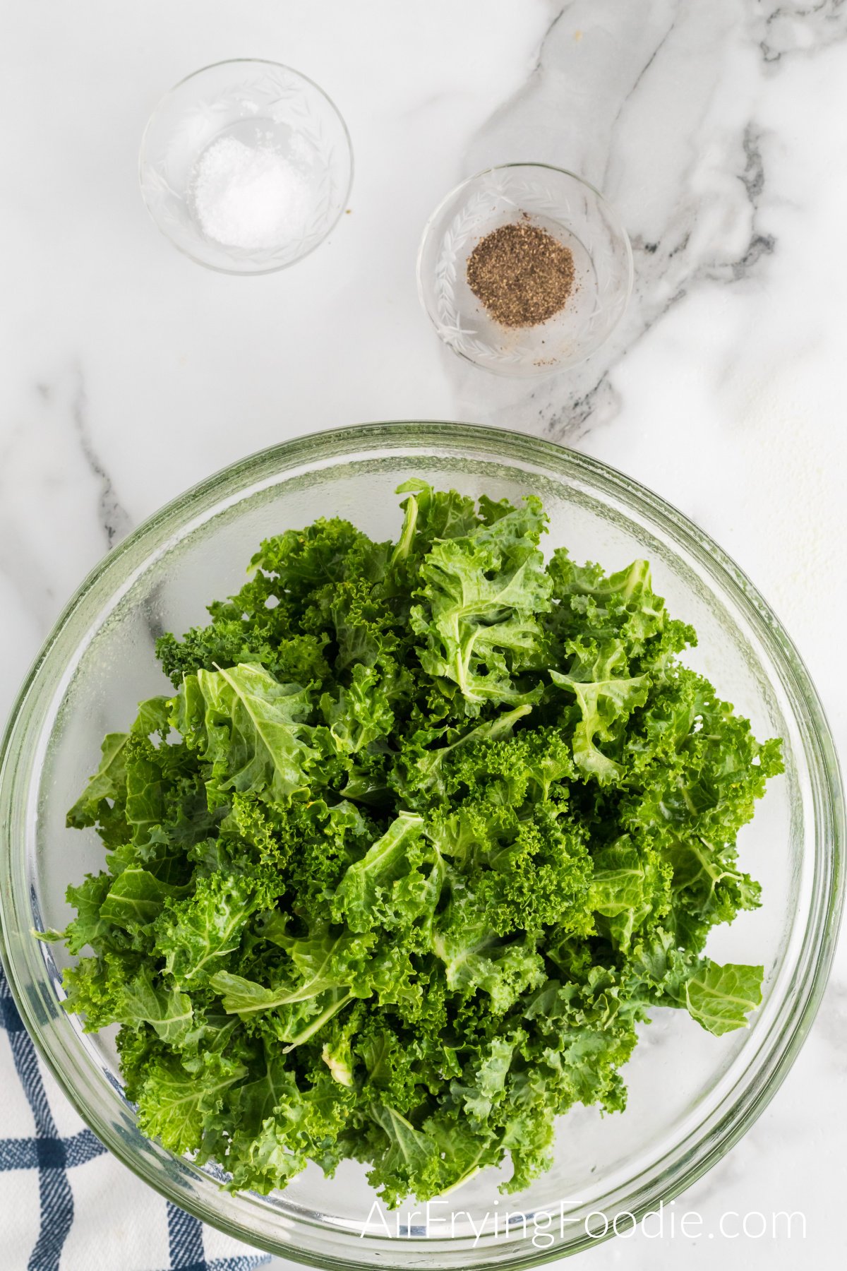 Picture of the kale that has been transferred to a glass bowl. Above the glass bowl of kale are two more glass bowls, one containing salt and the other containing pepper.