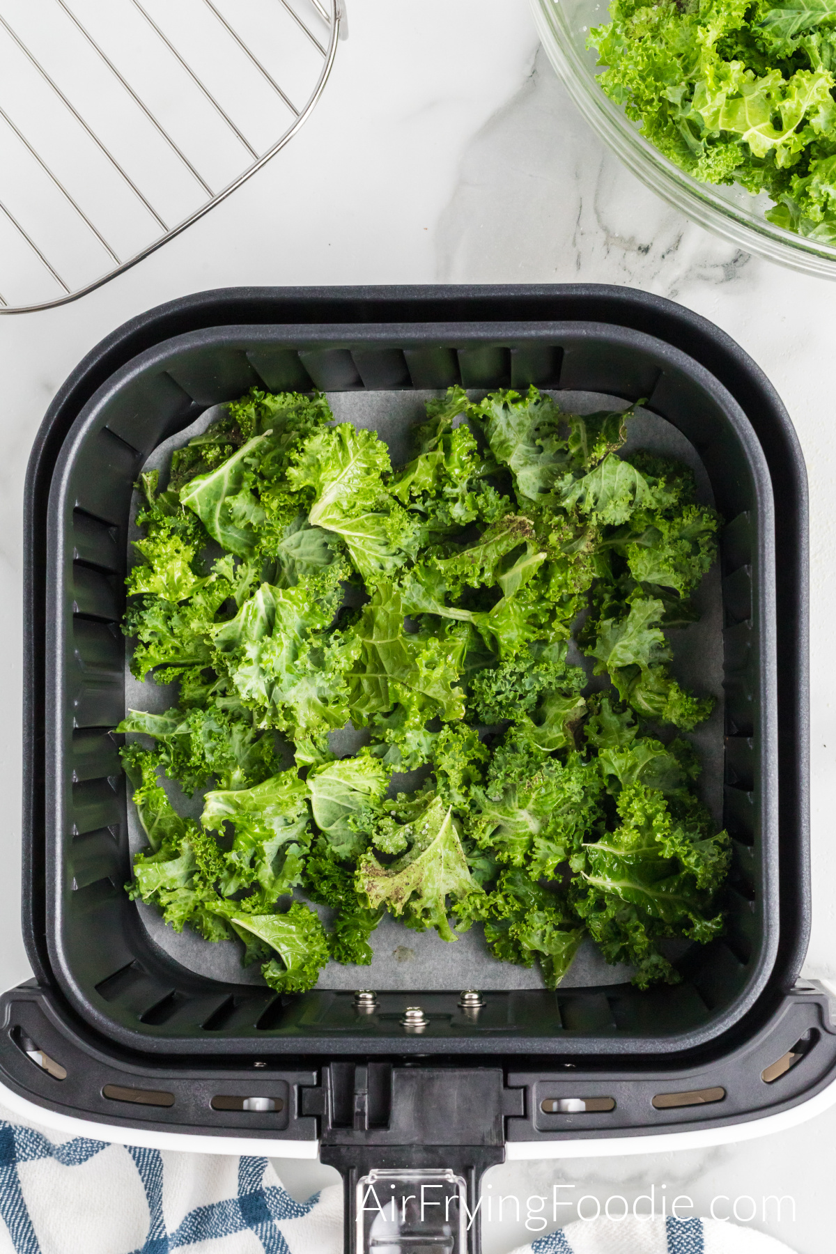 Seasoned kale that has been transferred to the air fryer basket and is ready to cook. The top right of the picture has a glass bowl with more kale leaves and a wire rack in the top right of the image.
