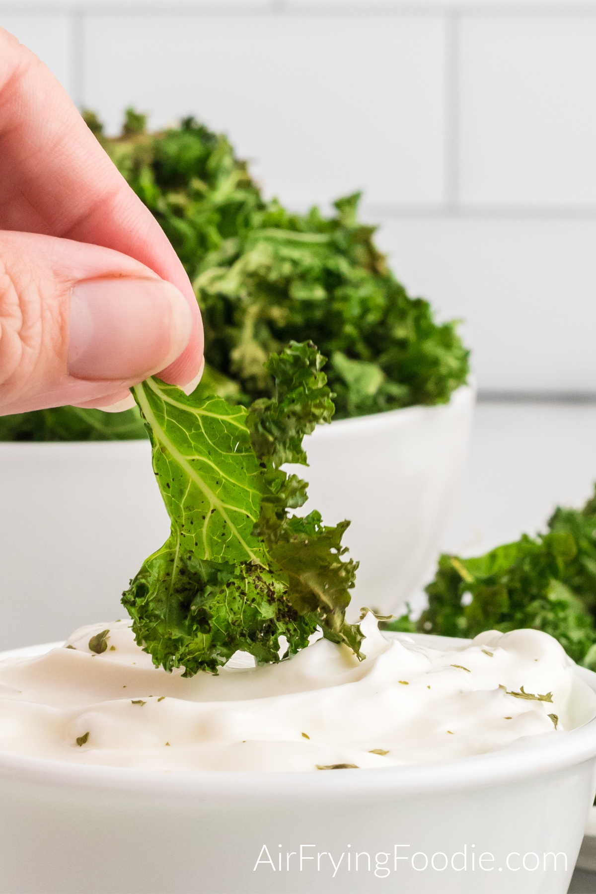 Close-up picture of kale chips cooked in the air fryer being dipped in greek yogurt. Behind the bowl of greek yogurt is a white bowl of kale chips. 