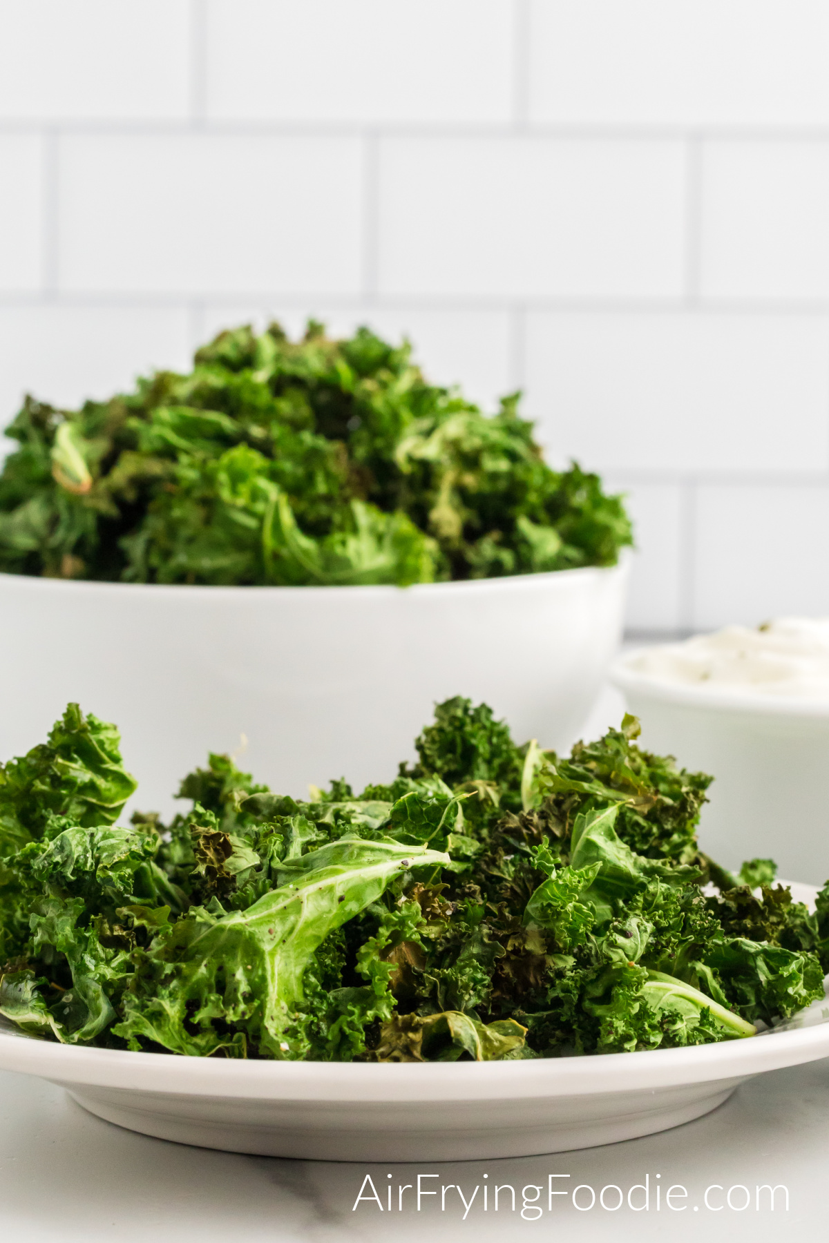 A close-up picture of kale chips cooked in the air fryer served on a small round white plate. Behind the small plate is a round bowl with more kale chips and a small round white bowl of yogurt dip to the right of the bowl. 