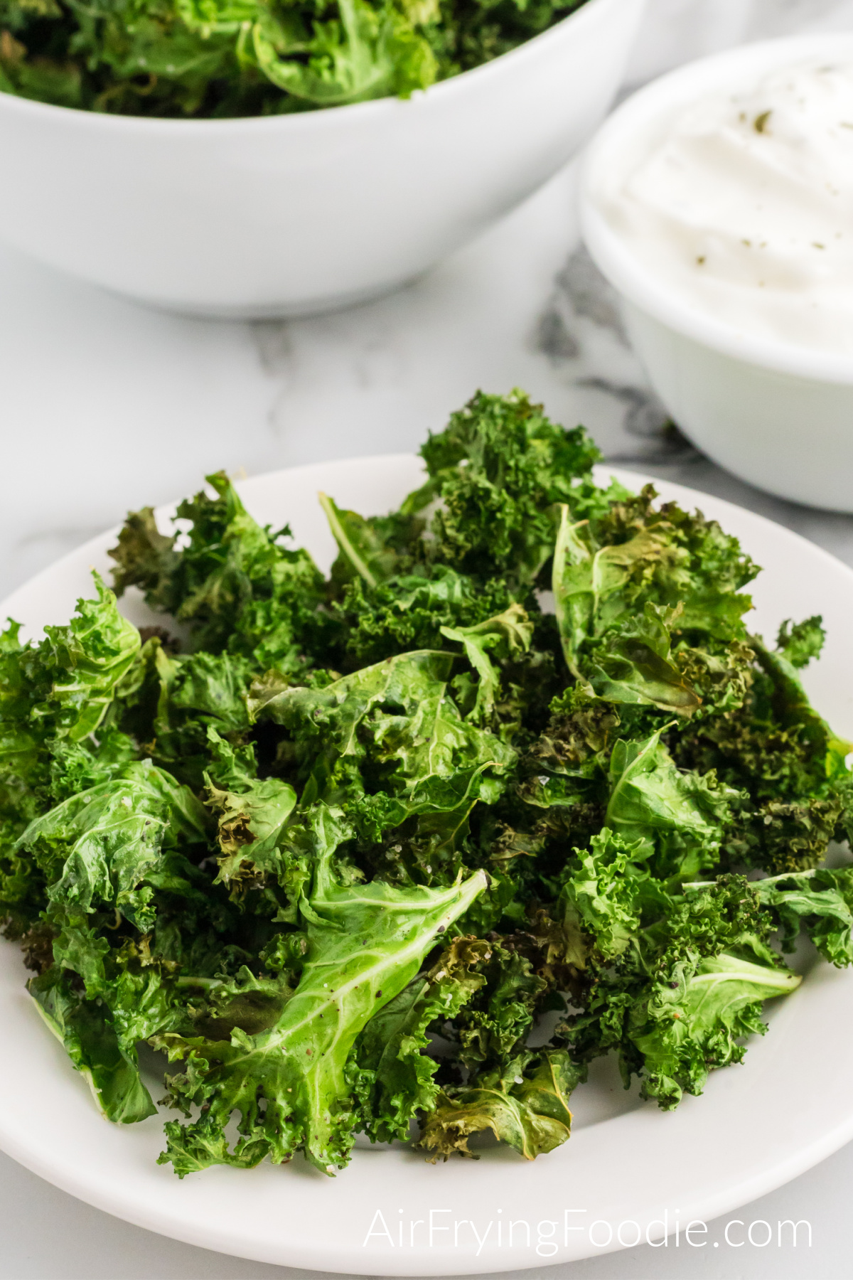 A round white plate of kale chips cooked in the air fryer. In the top left corner of the picture is a round white bowl with extra kale chips and a greek yogurt dipping sauce in a white bowl in the top right corner. 