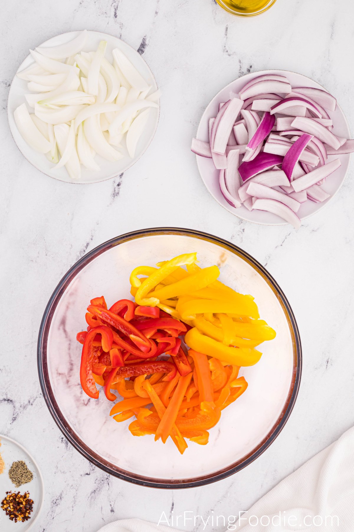 Overhead picture of all the peppers and onions sliced. There is one large round white plate with the peppers and two small round plates, one with sliced white onions and the second consisting of sliced red onions. A small portion of the seasoning plate is shown in the bottom right corner of the image, and a tiny bit of the glass bowl with olive oil is at the very top of the picture.