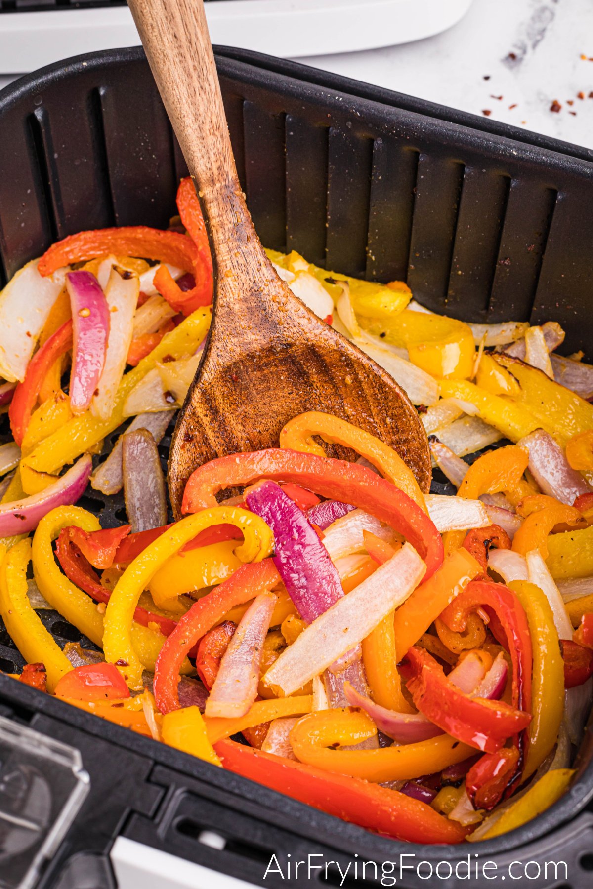 Close-up picture of peppers and onions in the air fryer basket with a wooden spoon stirring halfway through the cooking process.