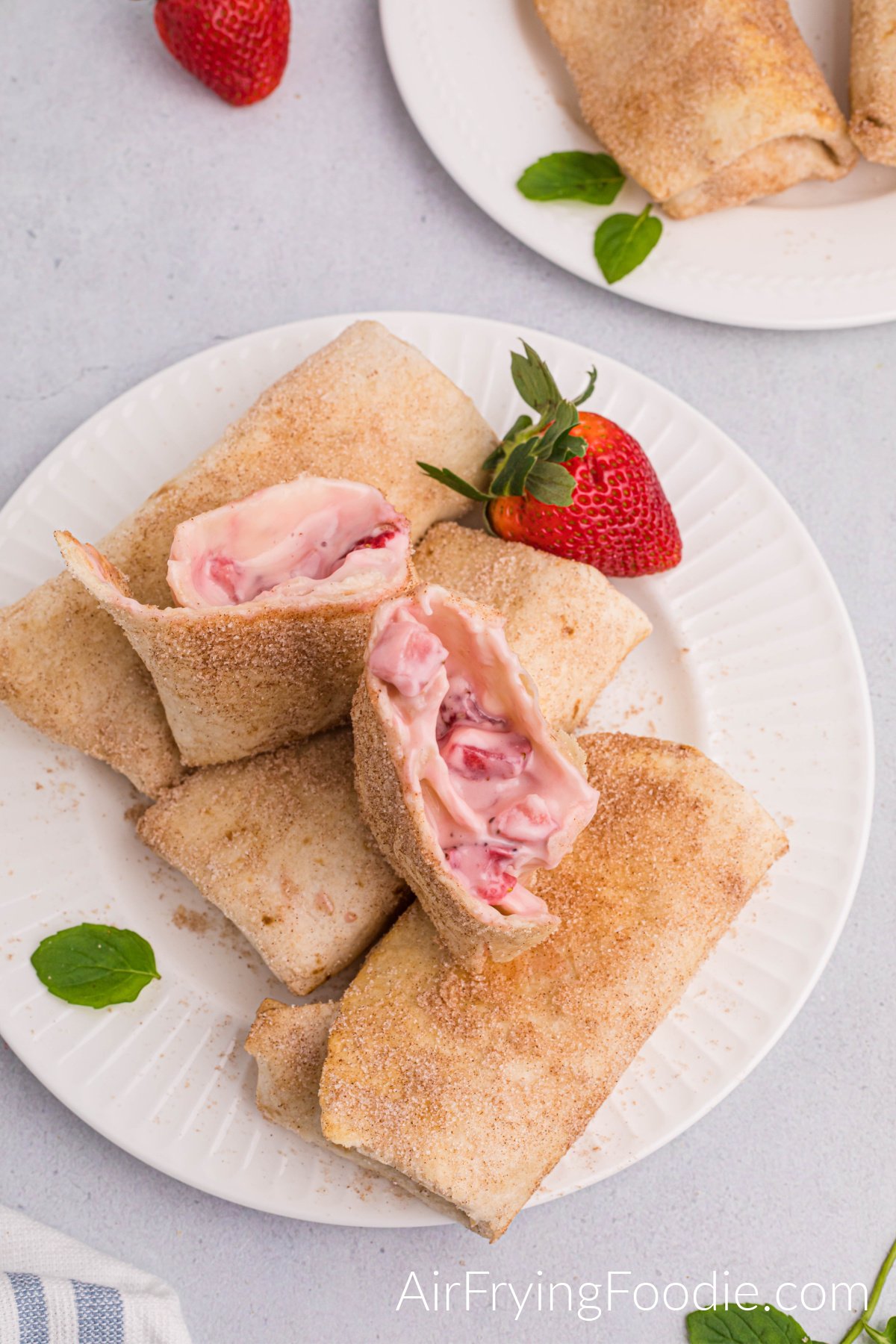 Overhead shot of three strawberry cheesecake chimichangas cooked in the air fryer and lined on a round white plate. A fourth chimichanga is cut in half, stands on top of the three chimichangas, and is garnished with a whole strawberry. In the top right of the picture is another round white plate with two chimichangas partially showing, and a whole strawberry is to the left. 