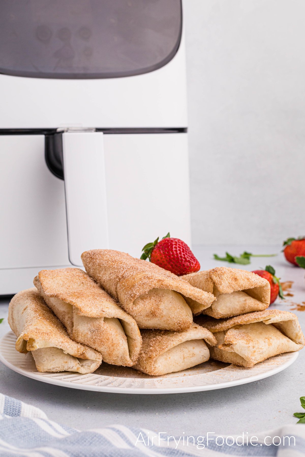 Round white plate with six strawberry cheesecake chimichangas cooked in the air fryer topped with cinnamon and sugar with a whole strawberry on top of the chimichangas. The base of the air fryer is behind the plate, and whole strawberries are on the table to the right. 
