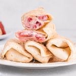 Air fryer strawberry cheesecake chimichangas served on a round white plate. There are three chimichangas lined on the bottom of the plate. One of the chimichangas is cut in half and stacked on top.