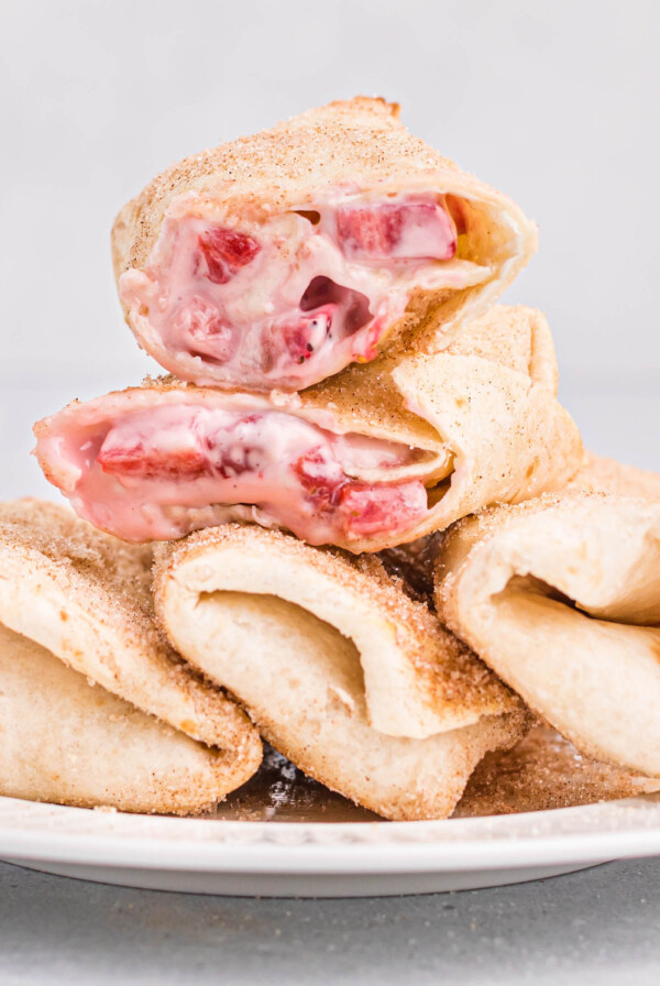 Air fryer strawberry cheesecake chimichangas served on a round white plate. There are three chimichangas lined on the bottom of the plate. One of the chimichangas is cut in half and stacked on top.