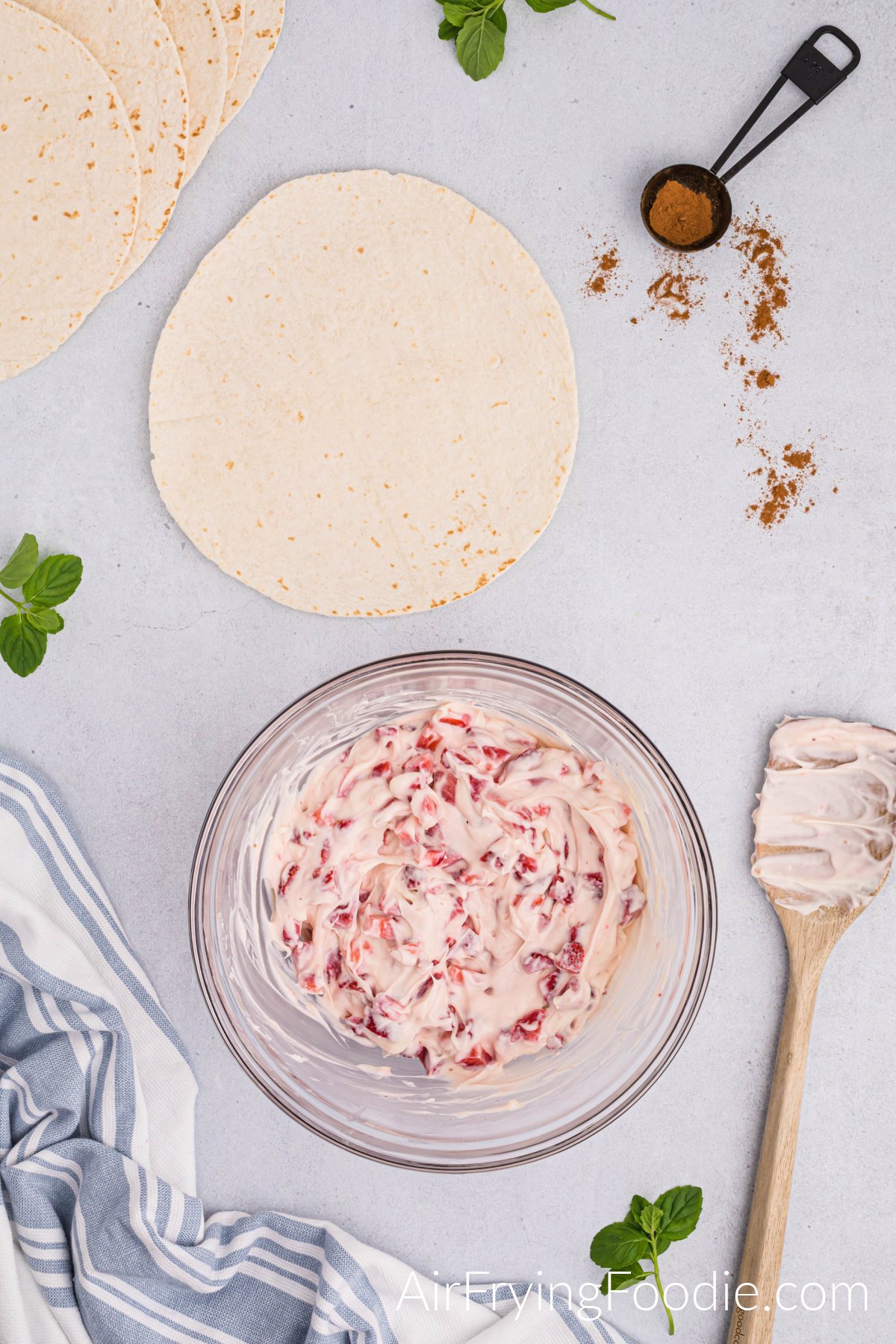 A glass bowl with the strawberries folded into the cream cheese and marshmallow mixture. There is a wooden spoon to the bowl's right with some mixtures on it. Above the bowl are tortillas and a measuring spoon of cinnamon.