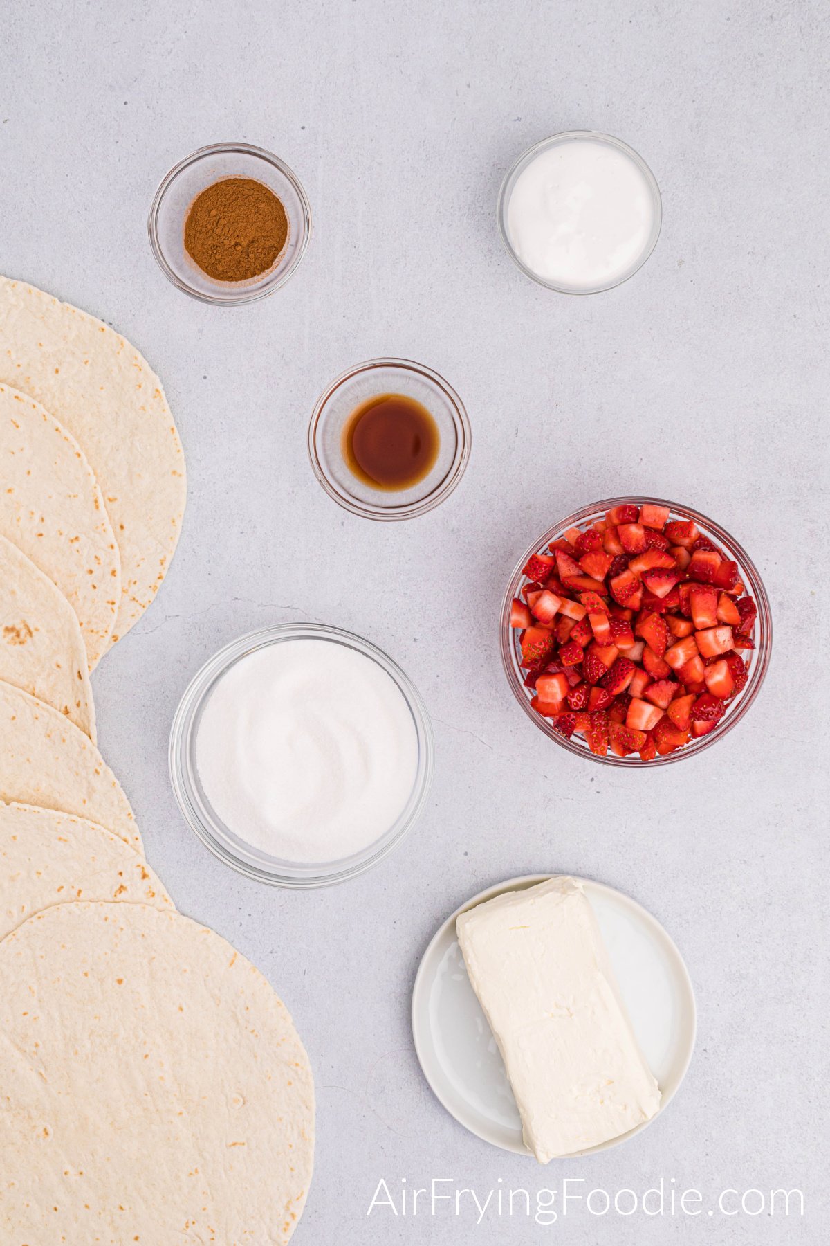 Ingredients shot with everything you need to make air fryer strawberry cheesecake chimichangas. To the left of the picture is a line of tortillas. The bottom of the image has a small rimmed white plate with a block of cream cheese. The rest of the photograph are bowls, each containing an ingredient; sugar, cut-up strawberries, vanilla extract, cinnamon, and marshmallow cream.
