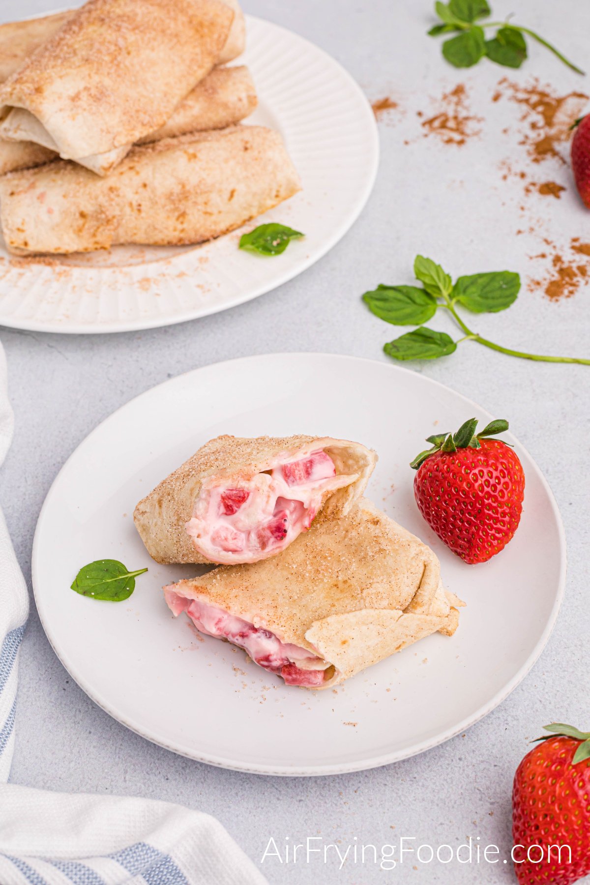 Air fryer strawberry cheesecake chimichanga cut in half, partially stacked and served on a round white plate with a whole strawberry and basil leaf as a garnish. A portion of a whole strawberry is in the bottom right corner of the picture. There is a round white plate in the top left of the photograph stacked with more strawberry chimichangas, and to the right is more basil garnish and a small portion of a strawberry showing. 