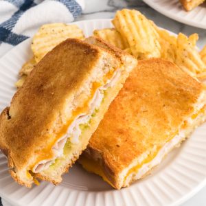 A picture of a turkey cheese melt after being cooked in the air fryer with cheese, mayo, and a side of chips was served on a round white plate.
