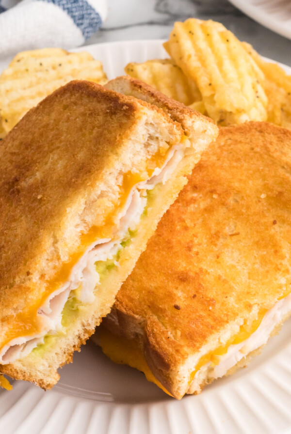 A picture of a turkey cheese melt after being cooked in the air fryer with cheese, mayo, and a side of chips was served on a round white plate.