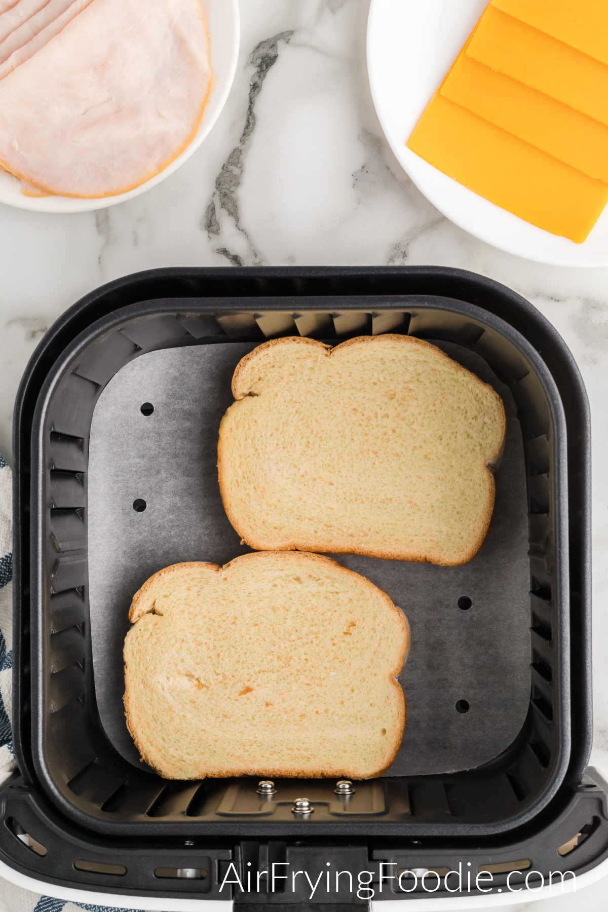 Air fryer basket with two slices of white bread with the butter side down. In the top left of the picture is a small white plate of turkey and a small white plate of cheese is in the top right of the image.