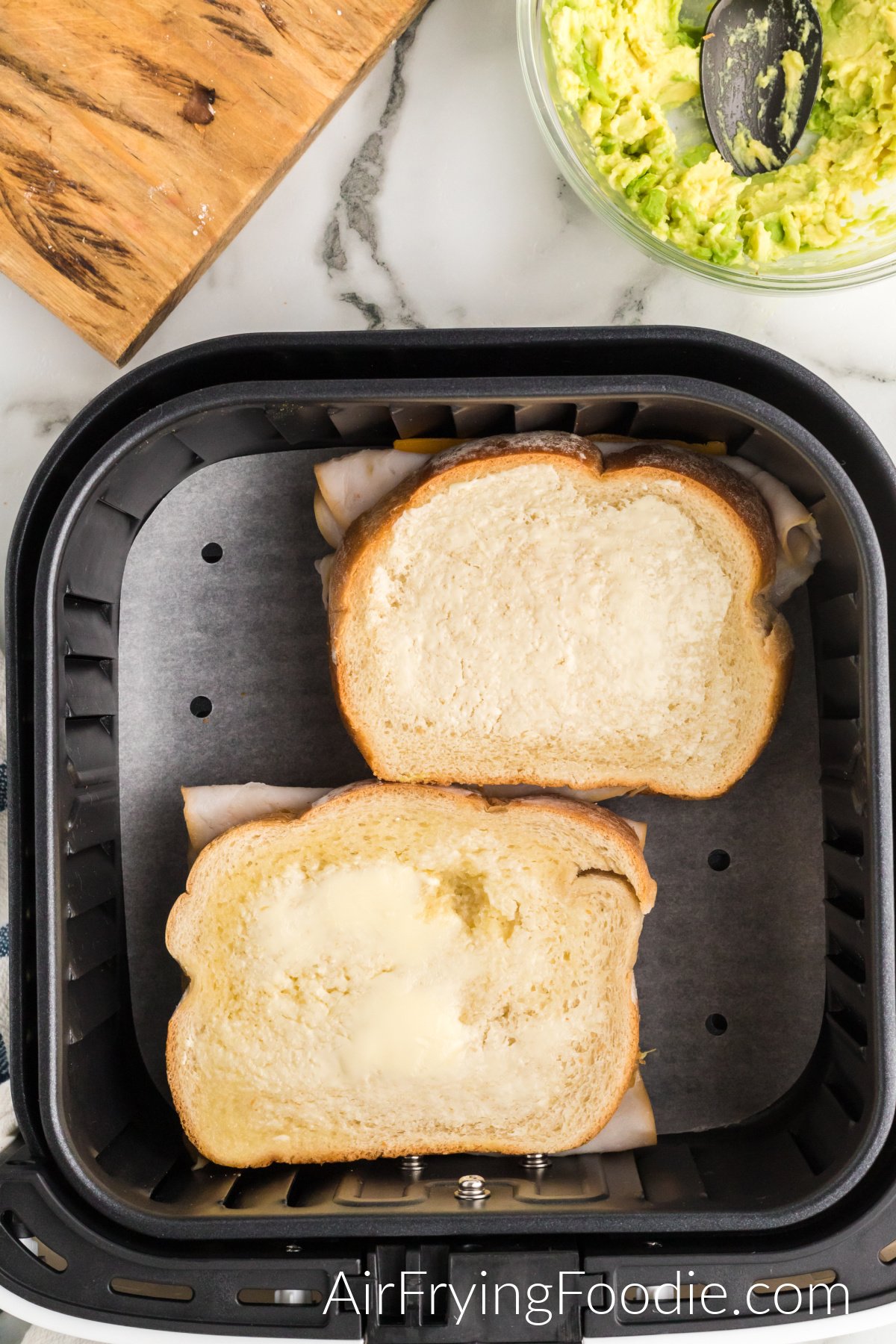 A picture of the air fryer basket with two turkey cheese melts with buttered sides up, ready to be cooked. There is a small portion of a cutting board in the top left of the picture and a glass bowl of mashed avocado in the top right.