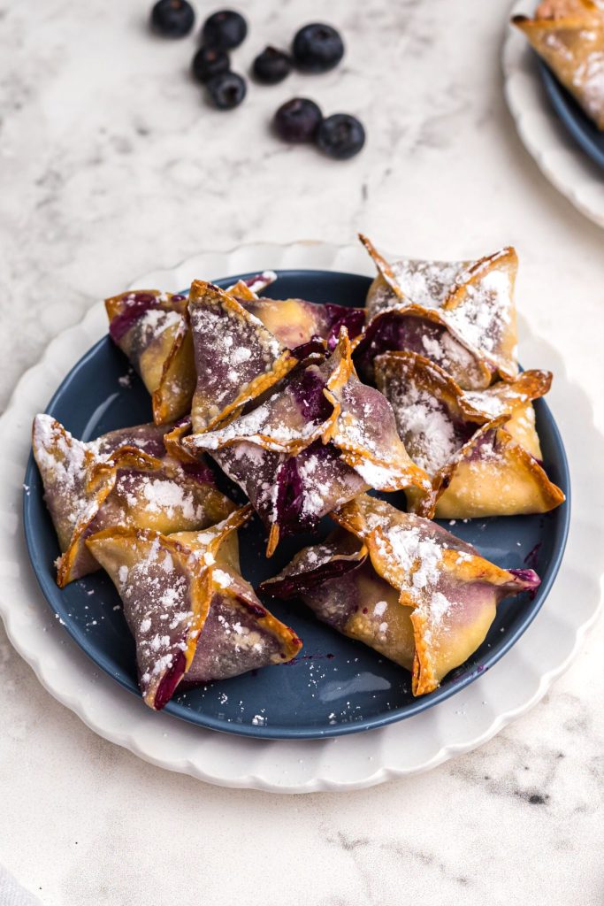Crispy wontons stacked on a blue plate sprinkled with powdered sugar. 