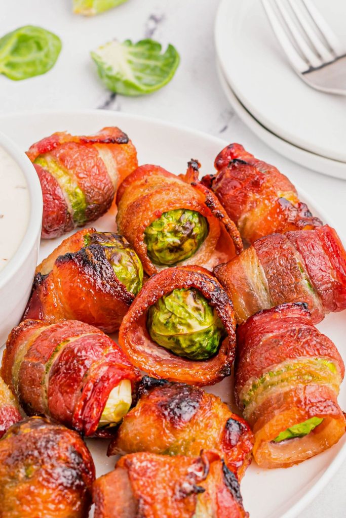 Golden juicy bacon wrapped brussels sprouts on a white plate with dipping sauce.