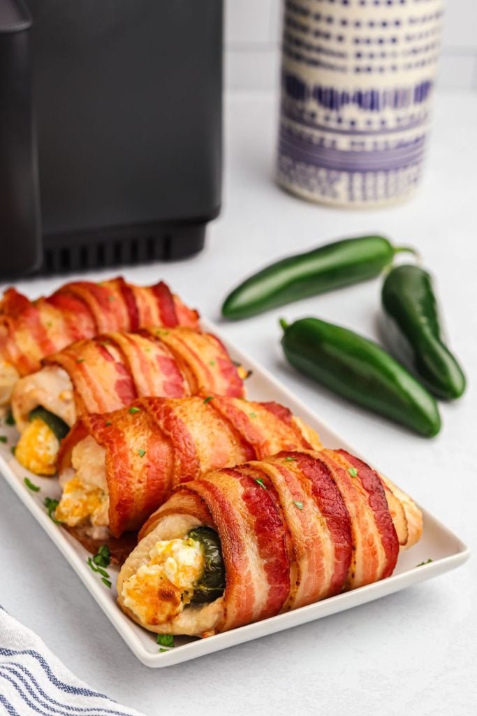 Crispy bacon wrapped chicken breast with jalapeno popper sticking out in the middle, on a white rectangle plate.