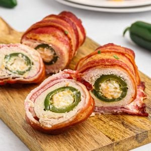 Crispy bacon wrapped around chicken, wrapped around jalapeno poppers on a wooden cutting board.