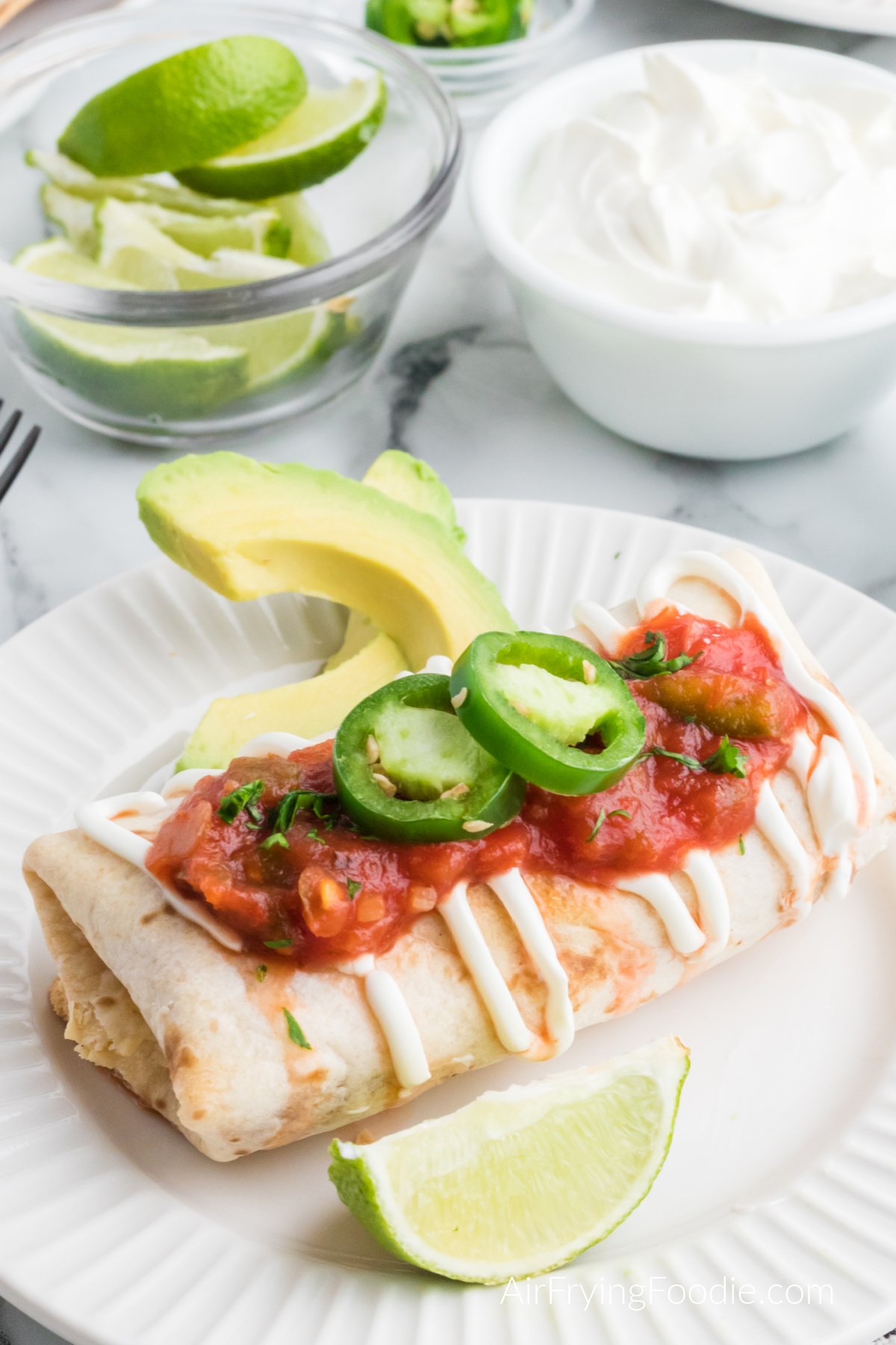 Chimichanga topped with sour cream, salsa, and sliced jalapenos on a white plate.