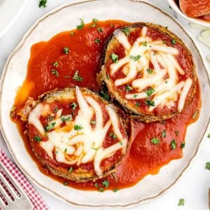 Crispy coated eggplant slices on a white plate covered in marinara sauce and melted cheese