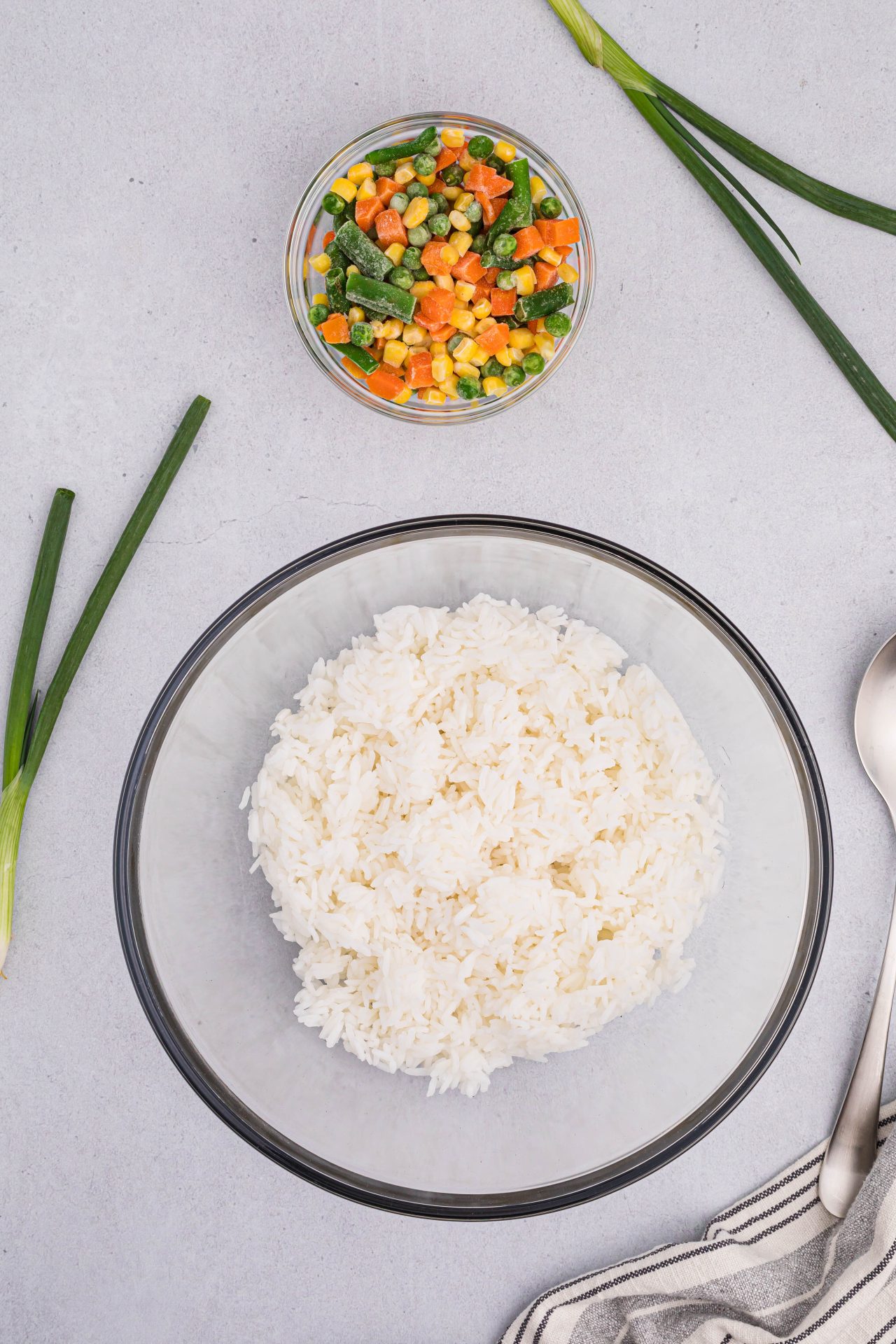 A large bowl of rice will be used to make air fryer fried rice. To the right of the bowl is a spoon, and a glass bowl of mixed vegetables is above the rice bowl.