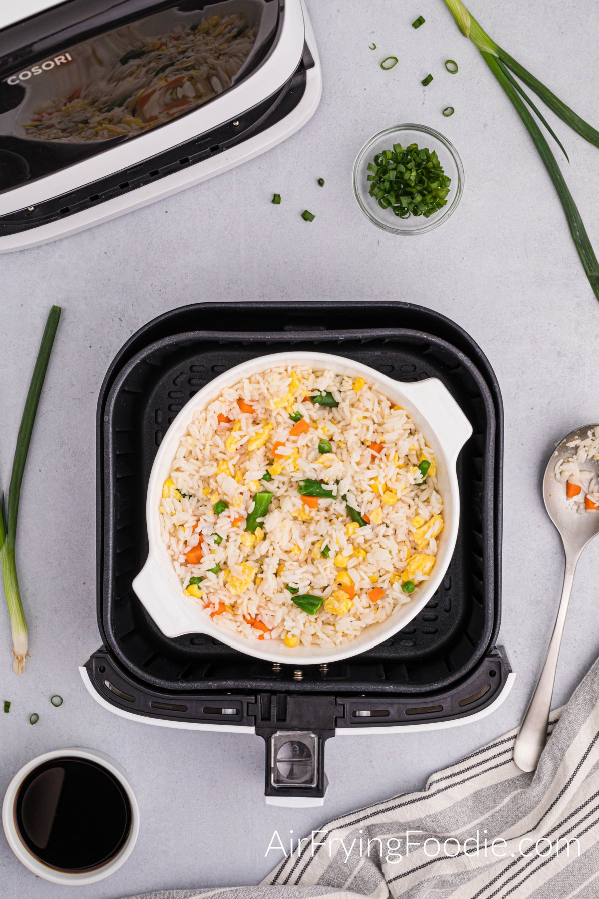 A large ramekin of fried rice in the air fryer basket with green onions sprinkled on top. There is a spoon to the right of the air fryer basket and a bowl of green onions above the air fryer basket. The base of the air fryer basket is at the top left of the picture.