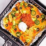 Crispy nachos in an air fryer basket topped with cheese, sour cream and salsa.