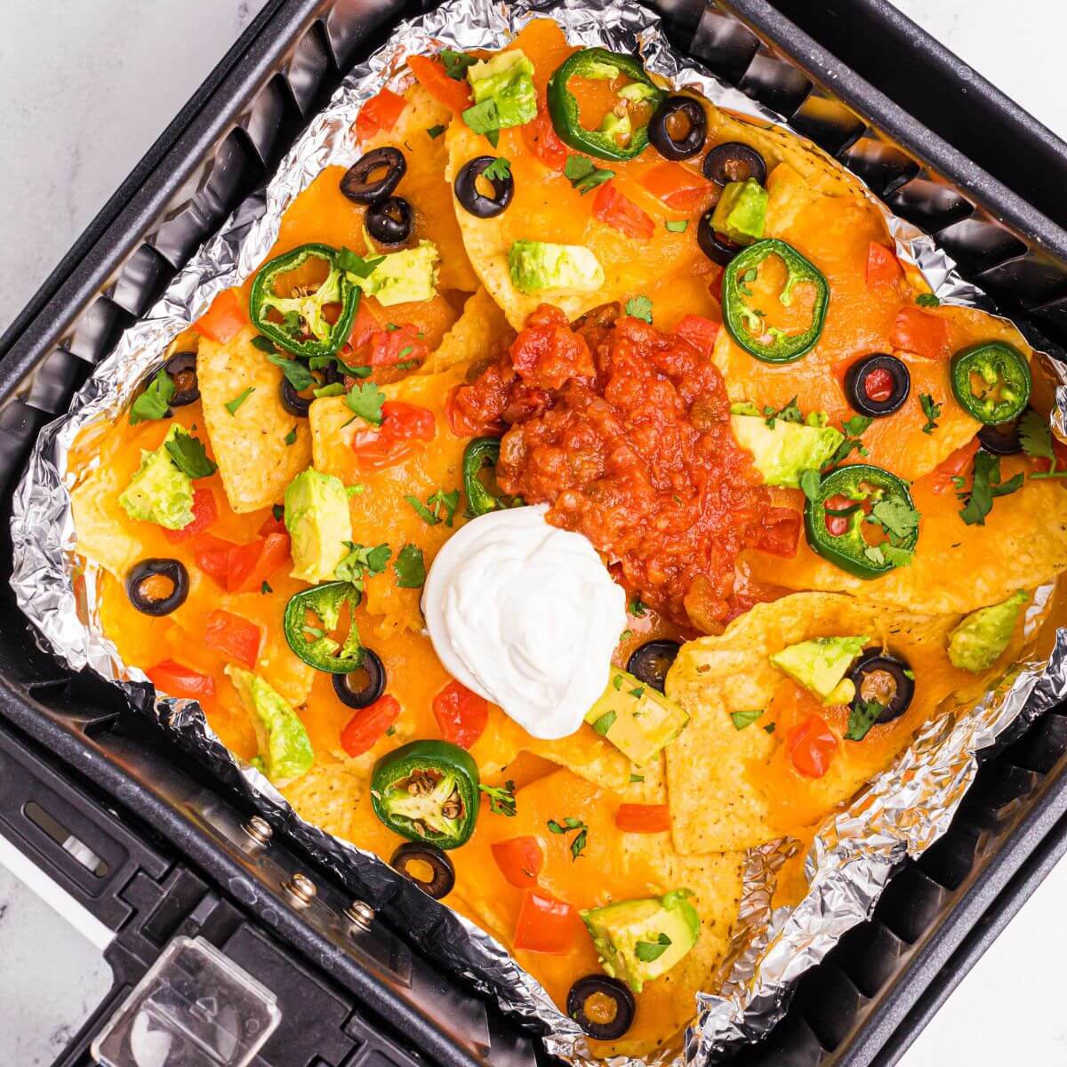 Crispy nachos in an air fryer basket topped with cheese, sour cream and salsa.