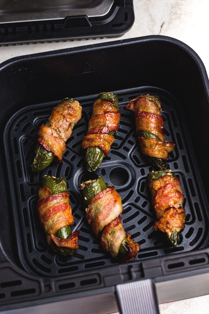 Crispy golden bacon wrapped around stuffed jalapenos in an air fryer basket. 