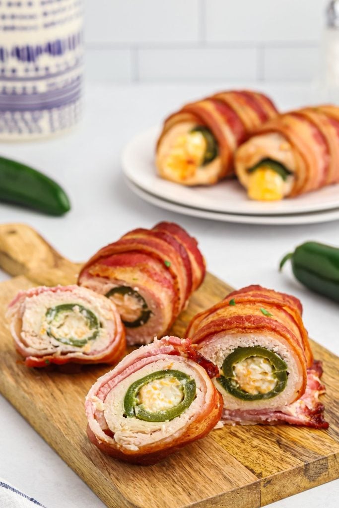 Crispy bacon wrapped around chicken and jalapenos stuffed with cheese on a wooden cutting board.
