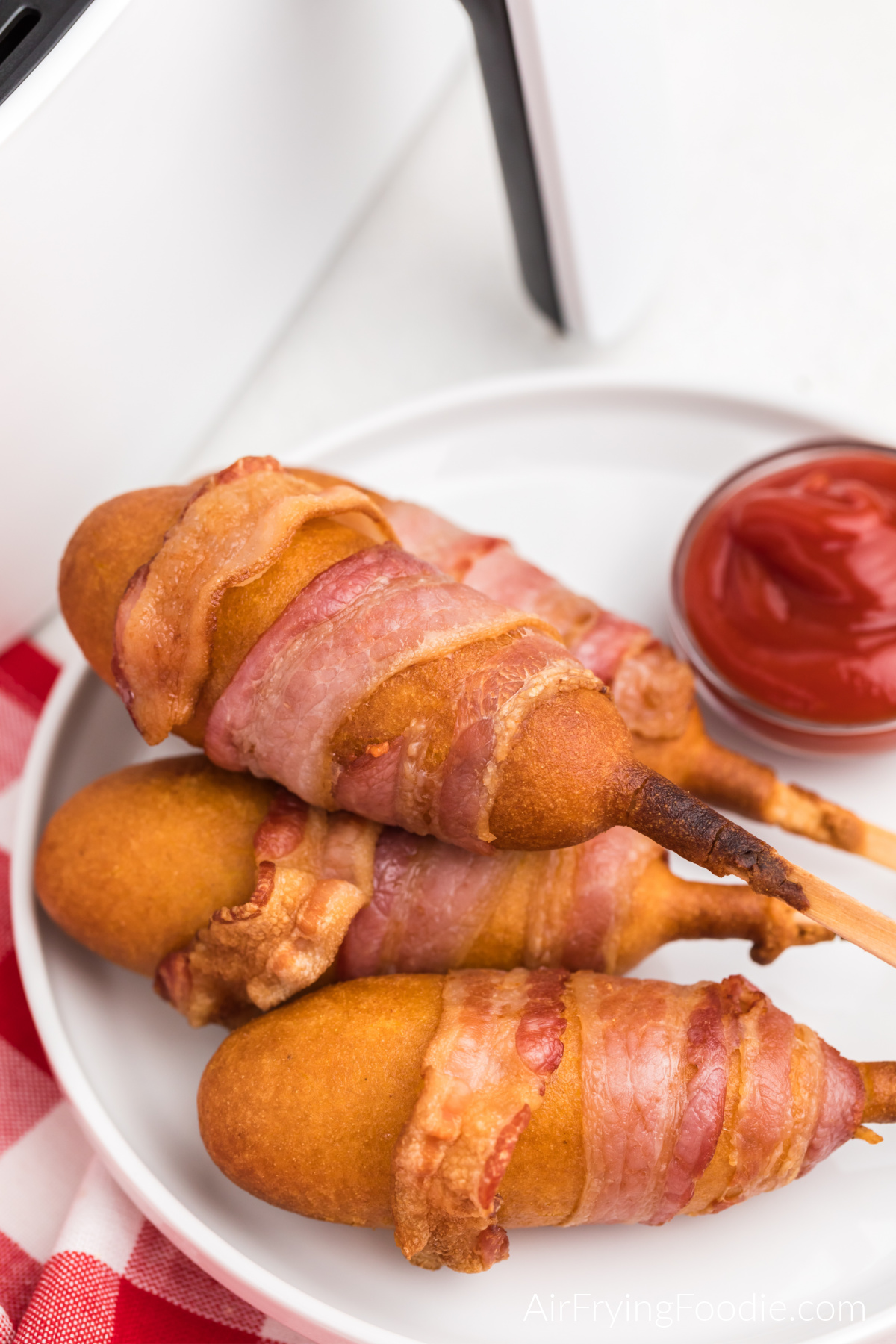 Bacon wrapped corn dogs on a white plate with a side of ketchup.