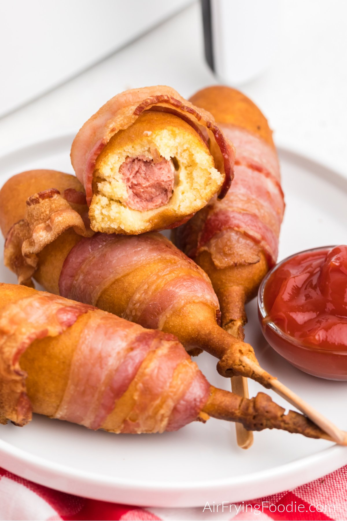 Bacon wrapped corn dogs stacked on a white plate and served with a side of ketchup. One air fried corn dog is missing a bite.