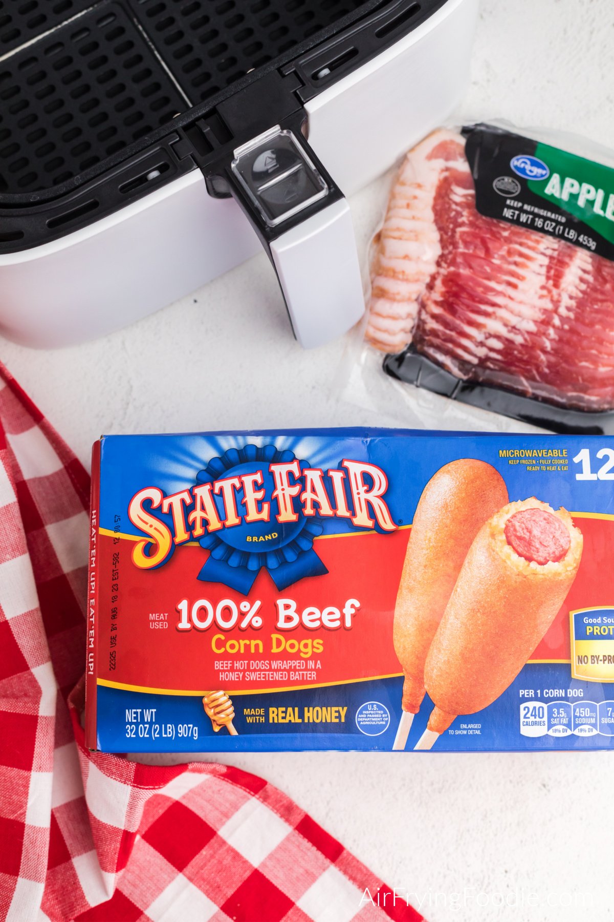 Package of bacon, frozen corn dogs, and basket of an air fryer on a white table.

