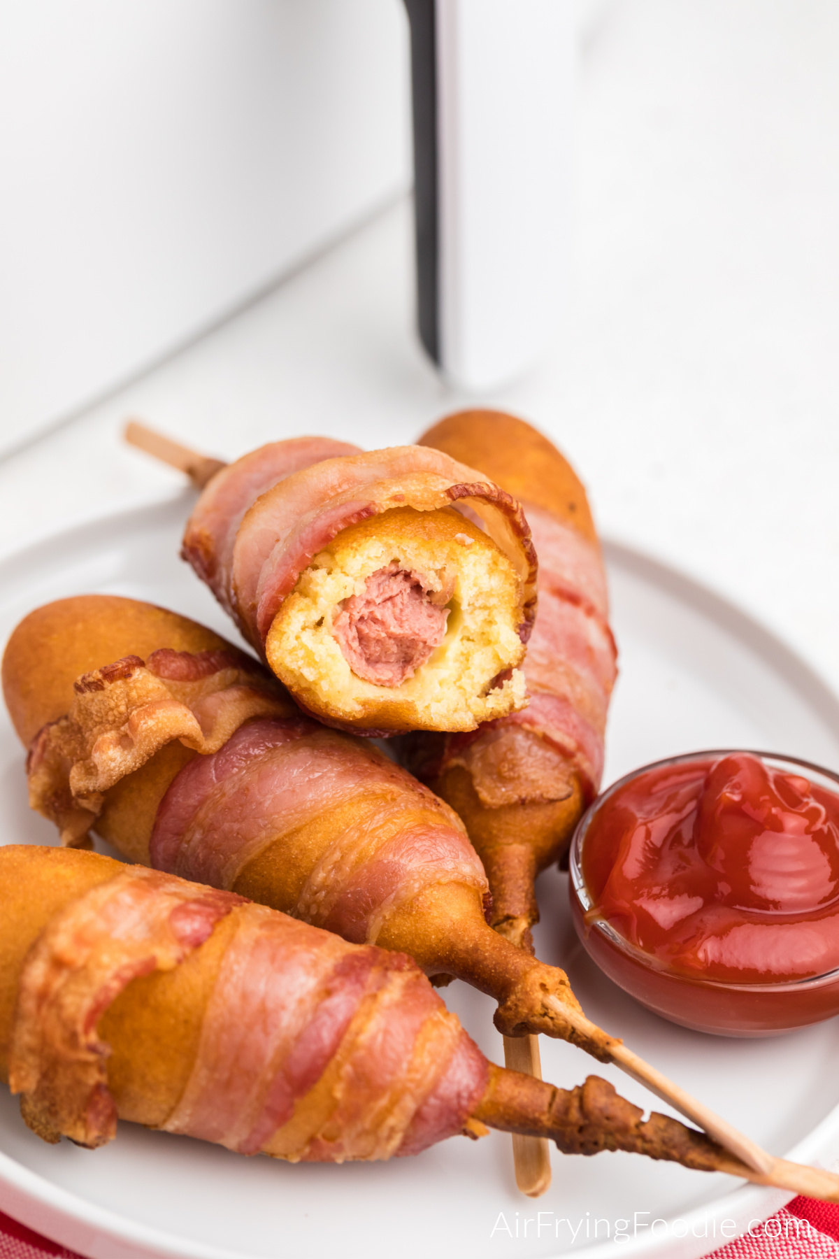 Bacon wrapped corn dogs made in the air fryer, stacked on a white plate with a side of ketchup, one missing a bite. 
