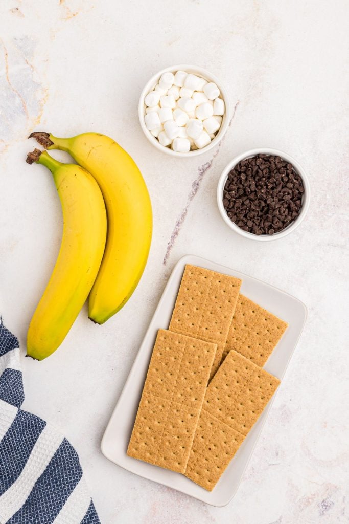 Graham crackers, bananas, chocolate chips and marshmallows measured on a marble table.