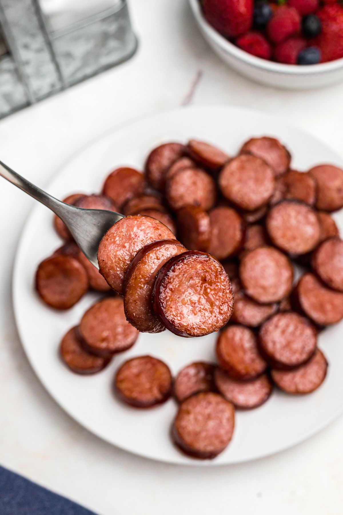 Easy Air Fryer Kielbasa 7 Fork holding pieces of kielbasa after being cooked in the air fryer over a white plate with more kielbasa, ready to eat.