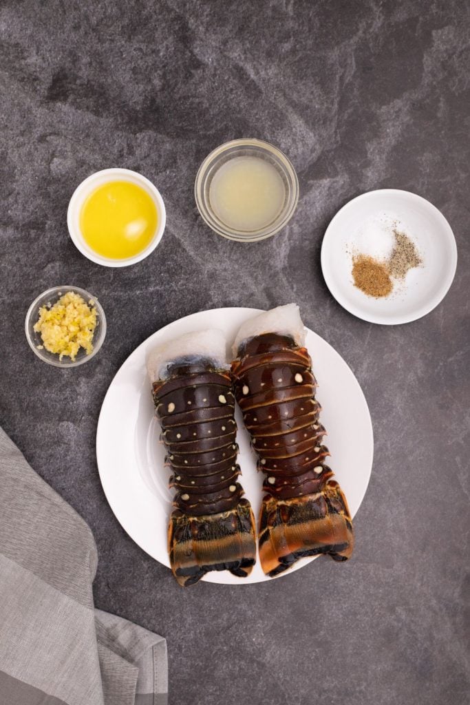 Lobster tails, melted butter, garlic, lemon juice, and seasonings, on a black marble table. 