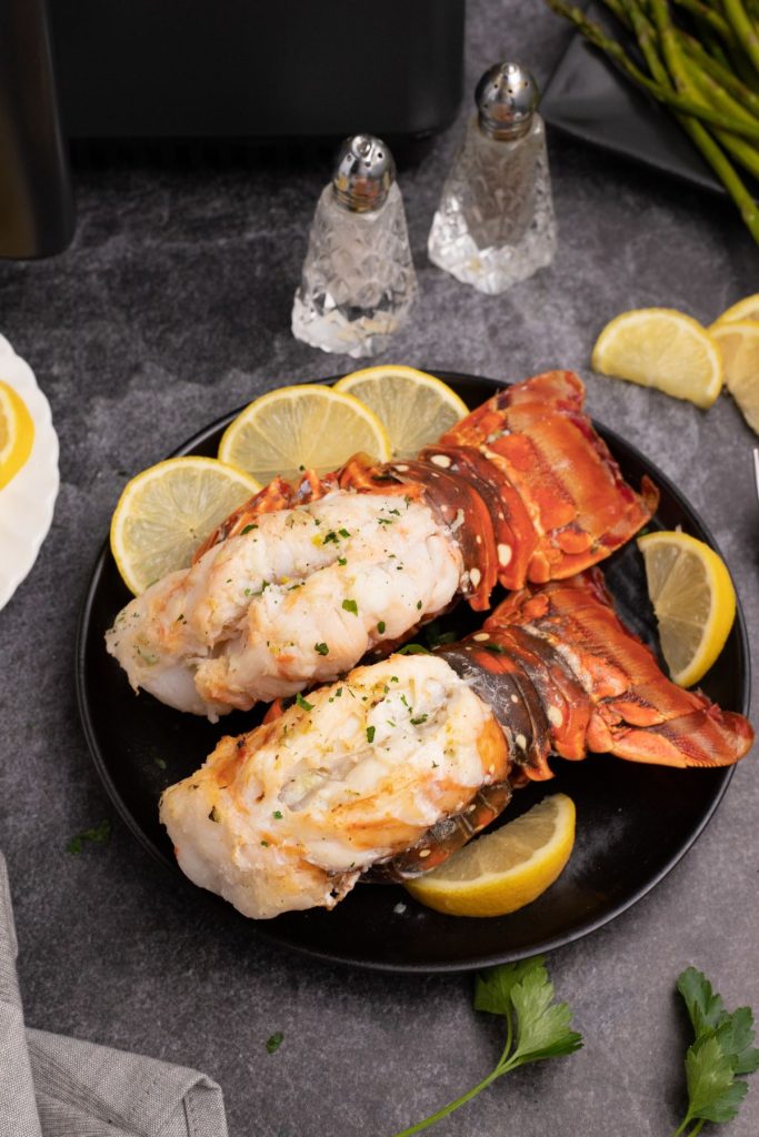 Juicy orange tailed lobster tails on a black plat with lemon wedges on a black marble table.