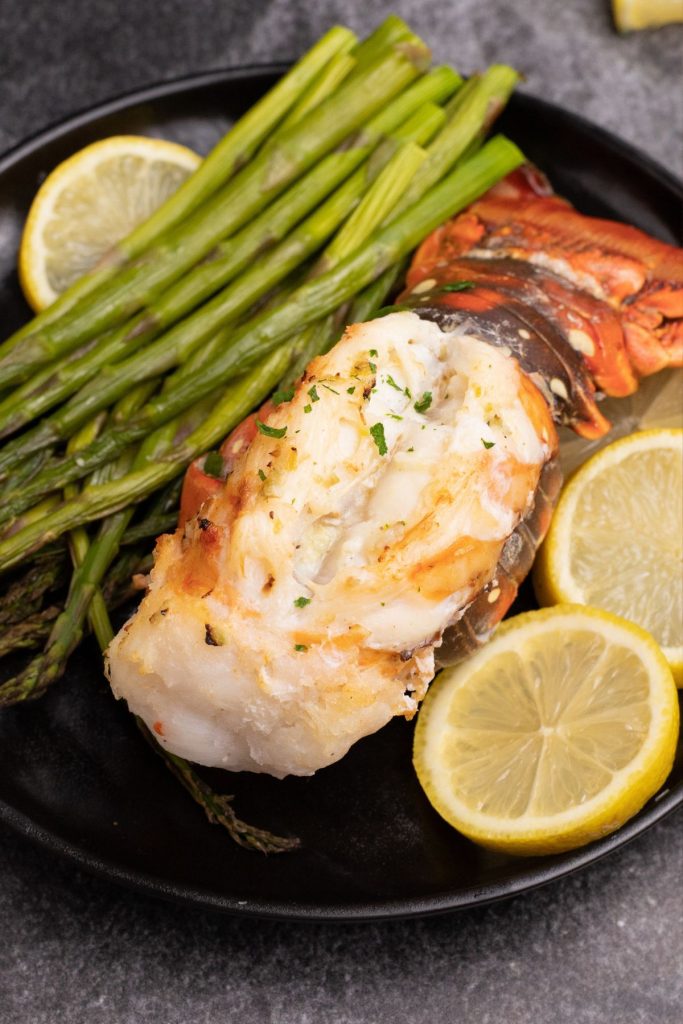 Lobster tail on a black plate served with asparagus and lemon wedges garnish.