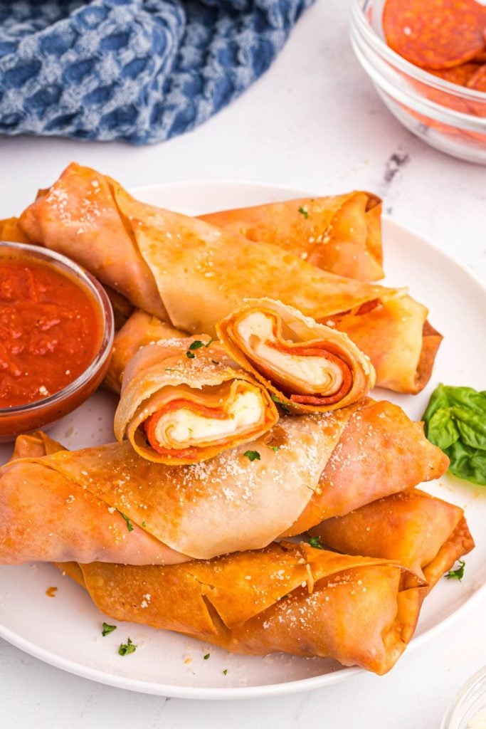 Golden crispy pizza filled egg rolls on a white plate with red dipping sauce. 