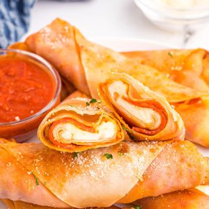 Golden crispy pizza egg rolls stacked on a white plate served with pizza sauce.