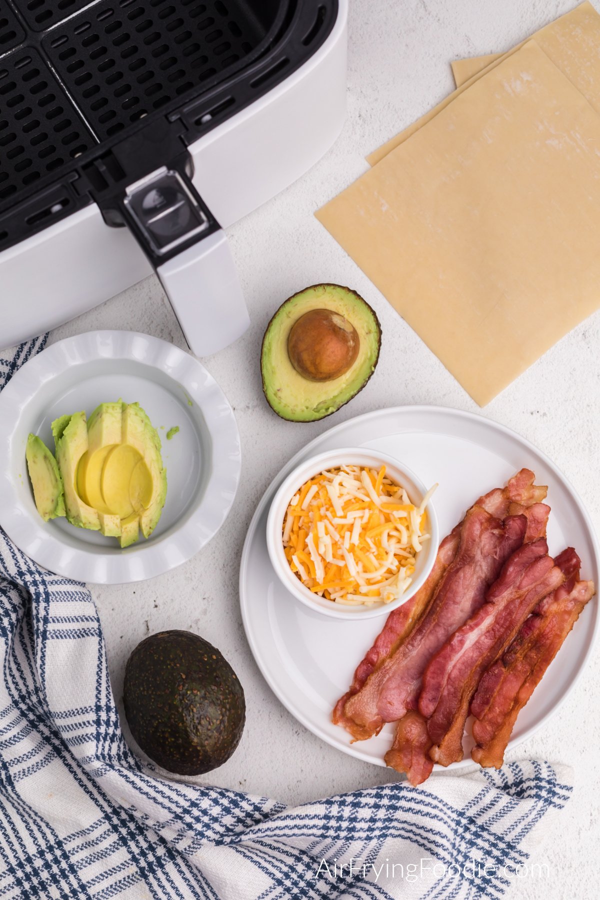 plate with cooked bacon, shredded cheese, bowl with sliced avocado, and avocados and egg roll wrappers in a table.