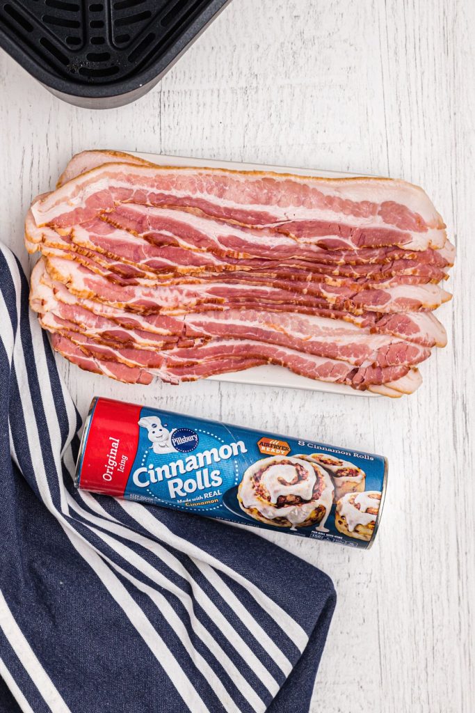 Uncooked bacon slices and a can of cinnamon rolls on a white wooden table.