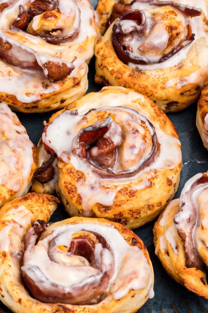 Close up photo of golden cinnamon roll wrapped around a slice of bacon and then drizzled with glaze frosting. 