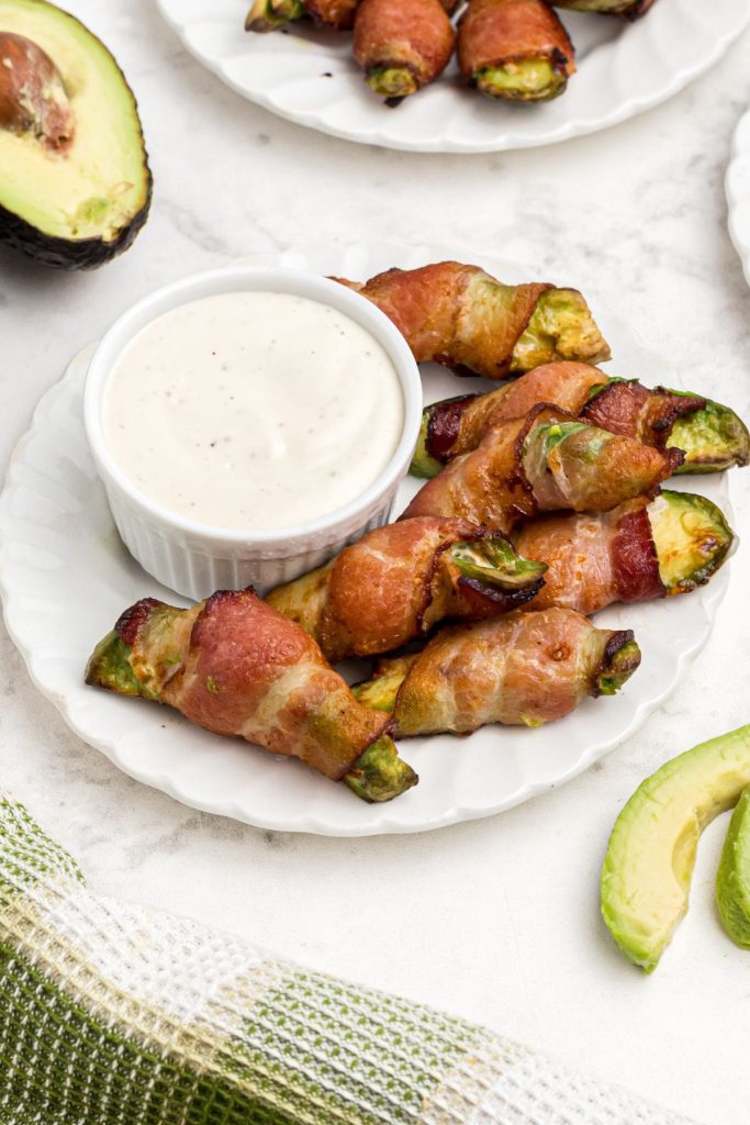 Crispy bacon wrapped around avocado wedges on a white plate with ranch dressing