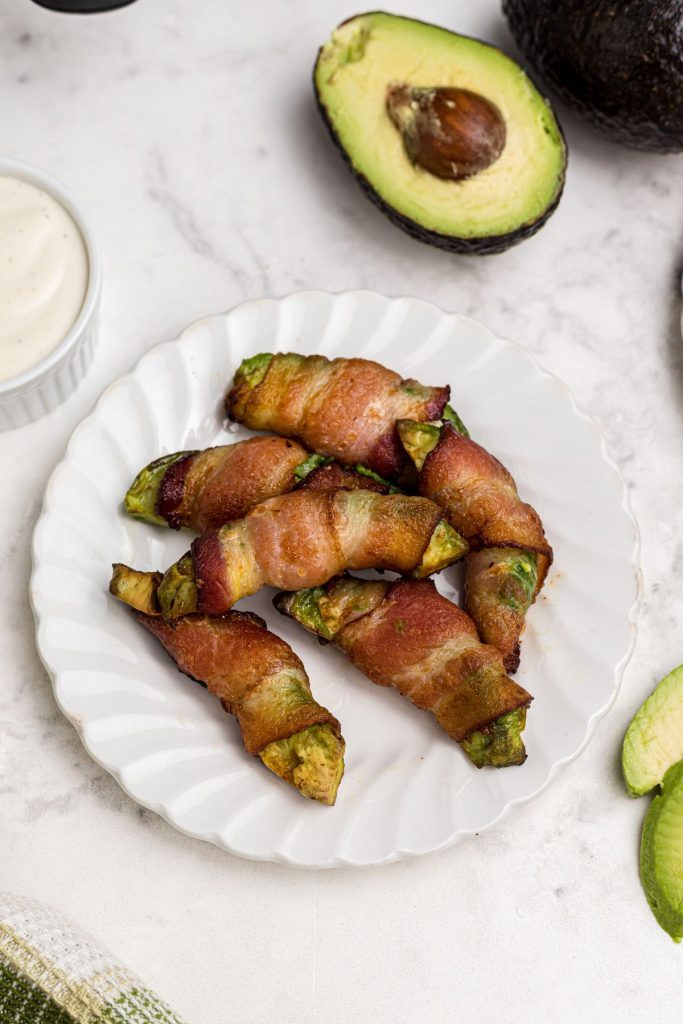 Crispy bacon wrapped around avocado wedge on a white plate on a marble table. 