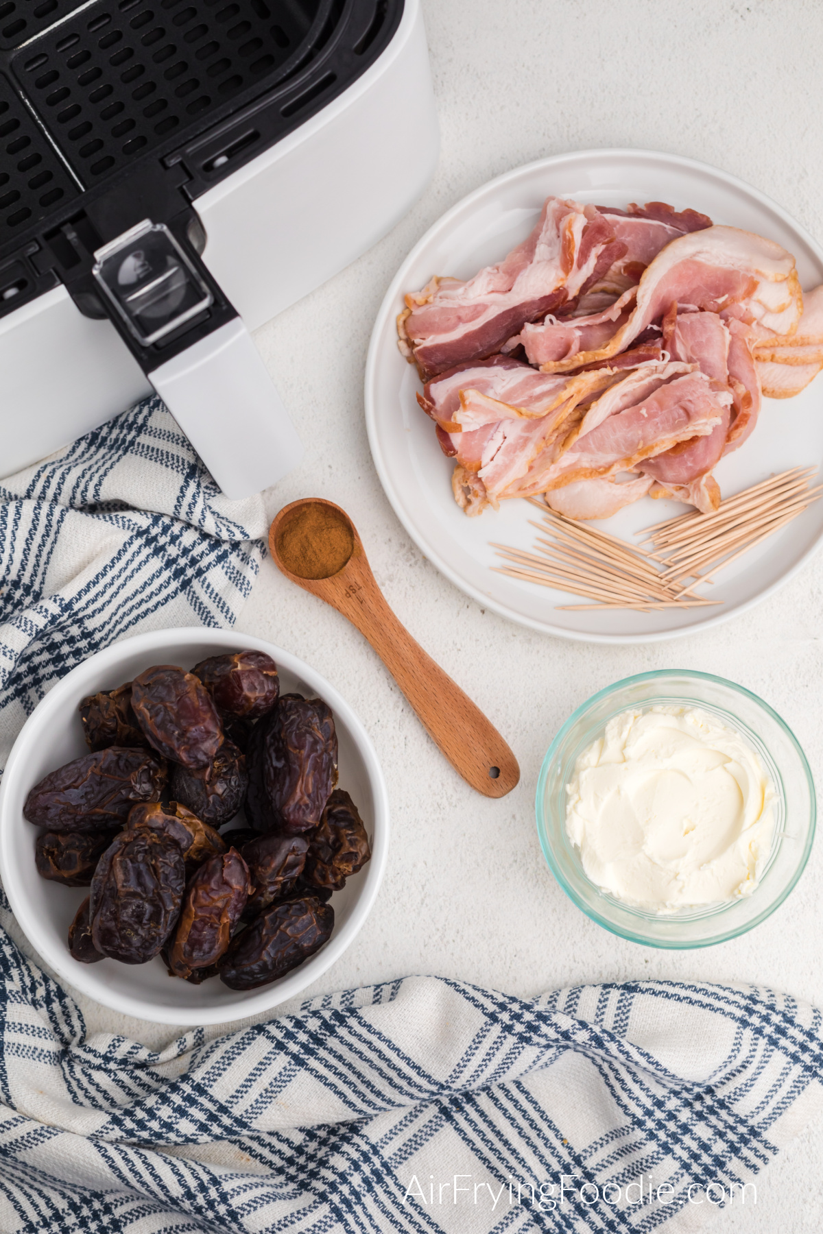 Table with air fryer basket, bowl of dates, cream cheese, plate with bacon and toothpicks, and a teaspoon of cinnamon.