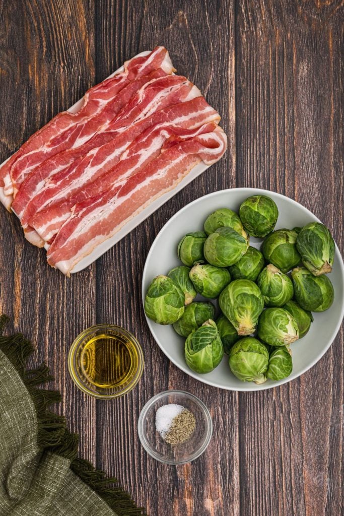 Brussels sprouts, bacon, olive oil, and seasonings on a wooden table. 