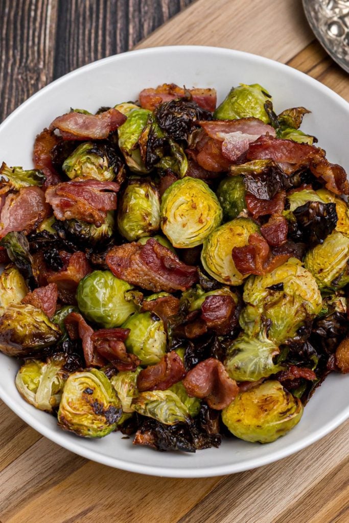 Crispy green cooked brussels sprouts tossed with crispy pieces of bacon in a white bowl on a wooden table. 