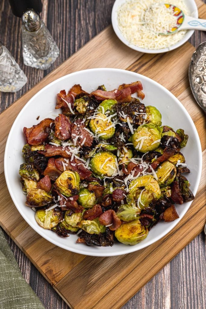 Green Brussels sprouts tossed with pieces of crispy bacon and shredded parmesan cheese in a white bowl.